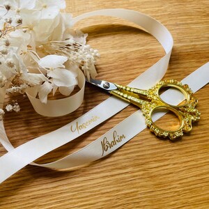 May include: White satin ribbon with the names "Yasemin" and "Ibrahim" written in gold lettering. A pair of gold scissors is lying on top of the ribbon.