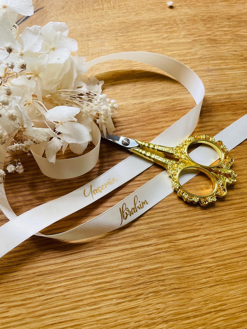 May include: White satin ribbon with the names "Yasemin" and "Ibrahim" written in gold lettering. A pair of gold scissors is lying on top of the ribbon.