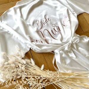May include: A white satin robe with the words "Bride Nur" in rose gold script. The robe has a tie closure and is decorated with floral accents. Dried flowers are arranged near the robe on a wooden surface.