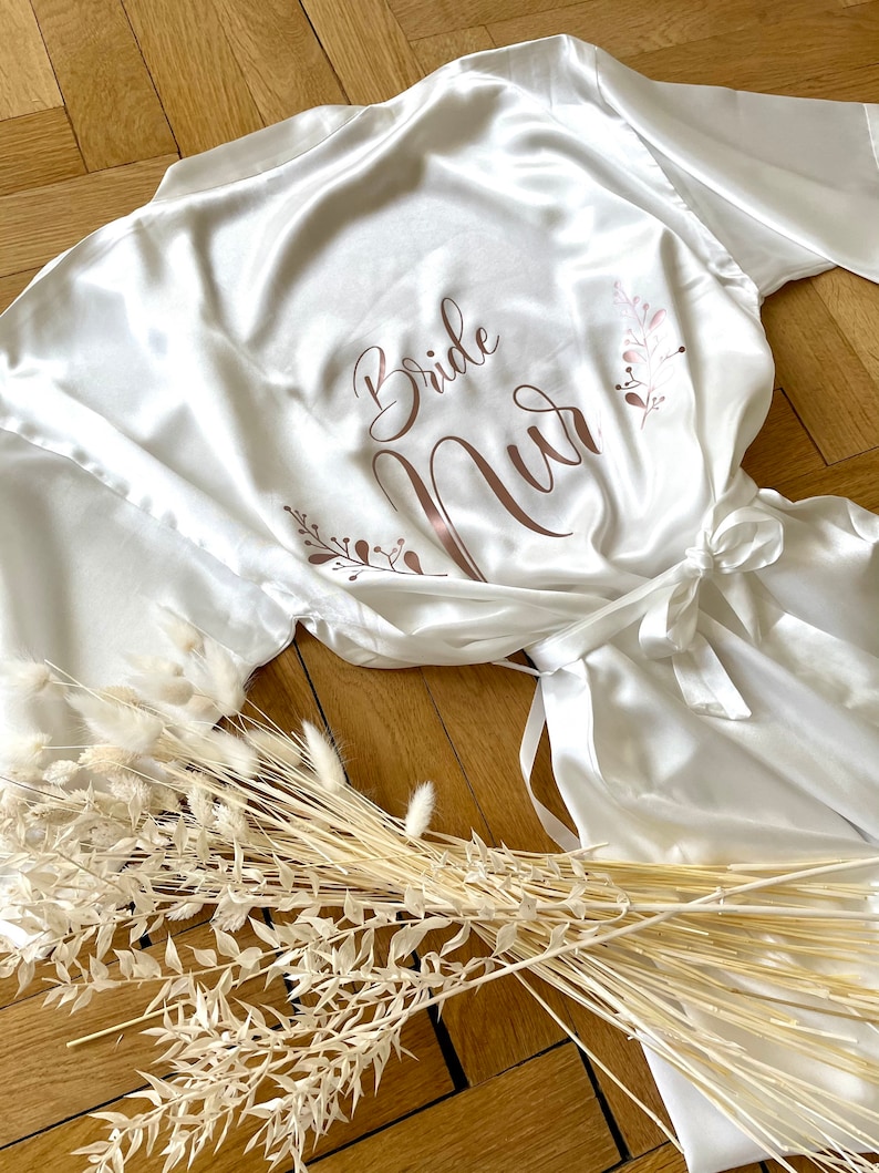 May include: A white satin robe with the words "Bride Nur" in rose gold script. The robe has a tie closure and is decorated with floral accents. Dried flowers are arranged near the robe on a wooden surface.