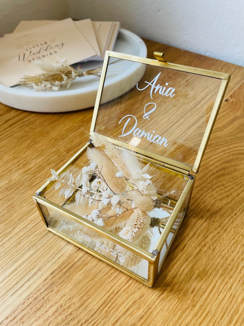 May include: A glass ring box with a gold frame and a clear lid. The lid has the names "Anya & Damian" written on it in white lettering. The box is filled with dried flowers and the date "27.08.2022" is written on the bottom of the box.