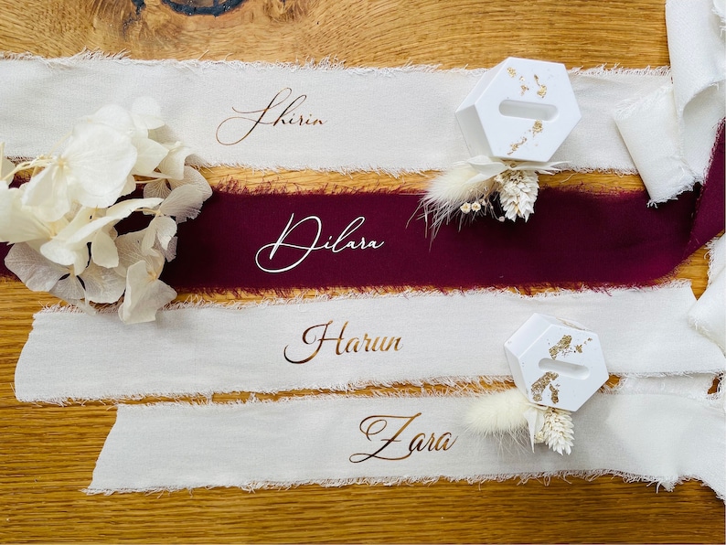 Op de afbeelding: Vier witte stoffen plaatskaarten met namen geschreven in kalligrafie. De namen zijn Shirin, Dilara, Harun en Zara. Elke plaatskaart heeft een kleine witte zeshoekige ringhouder met gouden accenten en een droogbloemenarrangement.