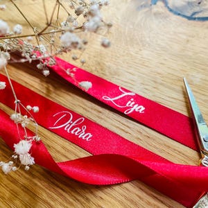 May include: Two red satin ribbons with the names "Dilara" and "Liya" printed in white cursive font. The ribbons are lying on a wooden surface with a pair of scissors in the background.