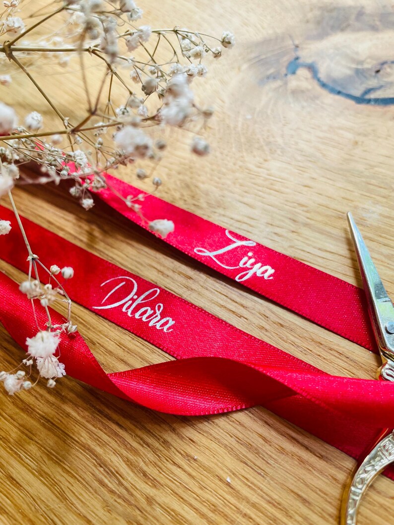 May include: Two red satin ribbons with the names "Dilara" and "Liya" printed in white cursive font. The ribbons are lying on a wooden surface with a pair of scissors in the background.
