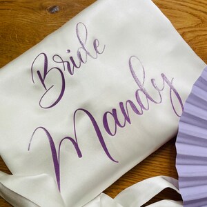 May include: Ivory satin robe with the words "Bride Mandy" in purple script. The robe has a matching satin tie and is partially visible. A lavender fan is in the background. The robe is likely for a wedding.