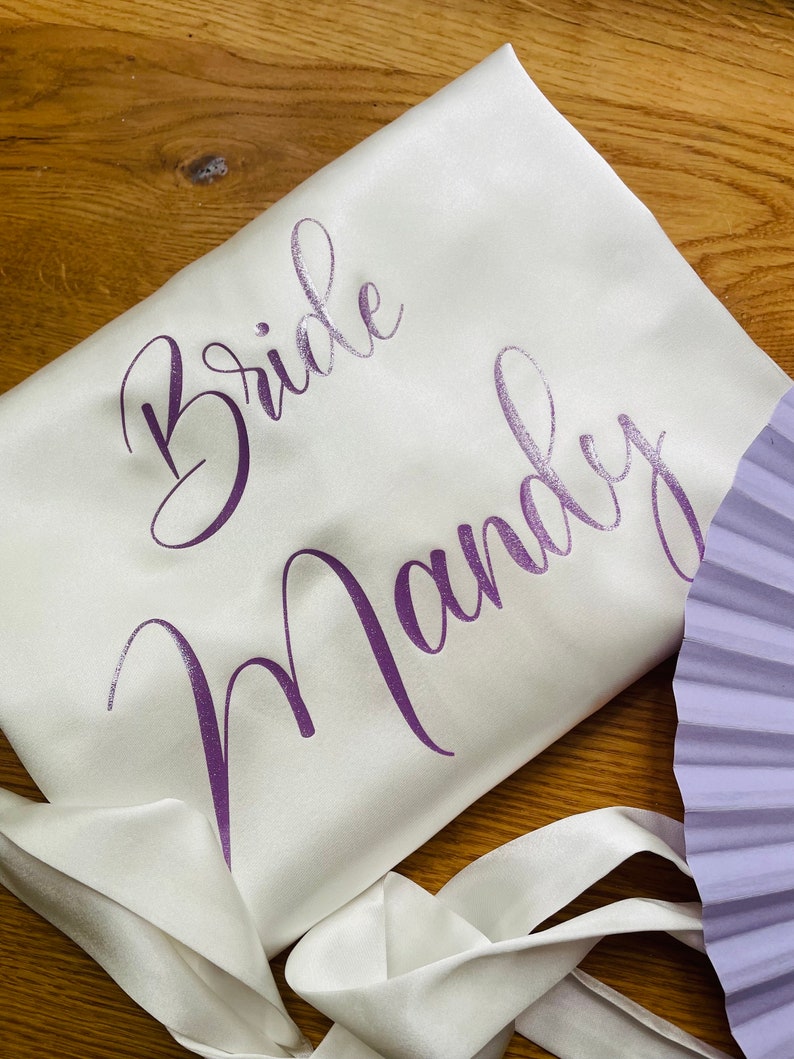 May include: Ivory satin robe with the words "Bride Mandy" in purple script. The robe has a matching satin tie and is partially visible. A lavender fan is in the background. The robe is likely for a wedding.
