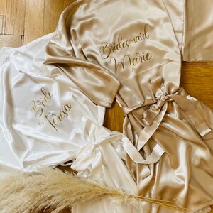 May include: Satin robes in ivory and champagne colors, with gold lettering. One robe says "Bride Teresa", and another says "Bridesmaid Marie". A bundle of dried pampas grass is placed near the robes. The robes have matching tie belts.