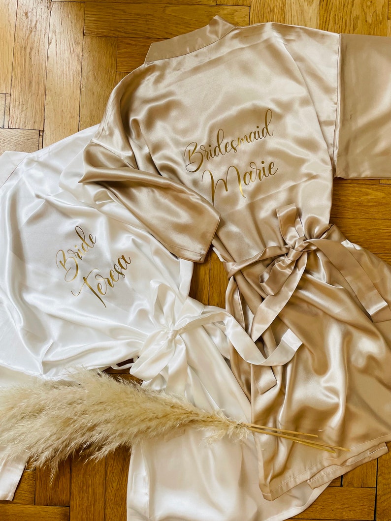 May include: Satin robes in ivory and champagne colors, with gold lettering. One robe says "Bride Teresa", and another says "Bridesmaid Marie". A bundle of dried pampas grass is placed near the robes. The robes have matching tie belts.