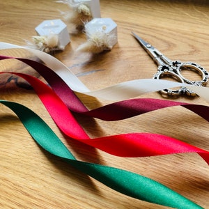 May include: Four spools of ribbon in red, burgundy, white, and green. The ribbon is arranged in a wave pattern on a wooden surface. A pair of silver scissors is also visible.