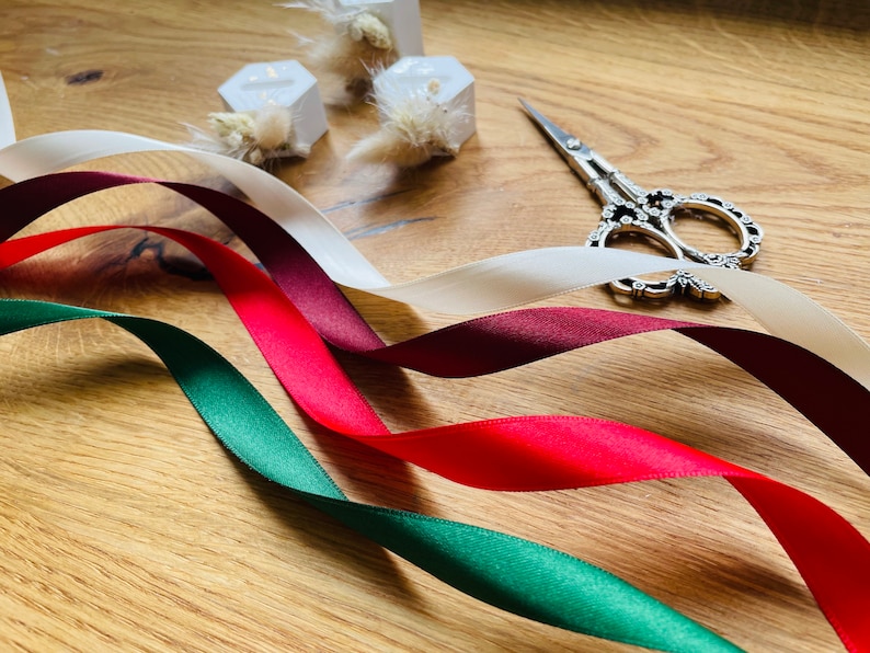 May include: Four spools of ribbon in red, burgundy, white, and green. The ribbon is arranged in a wave pattern on a wooden surface. A pair of silver scissors is also visible.