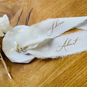 Op de afbeelding: Twee witte stoffen linten met gouden letters, een met de naam "Shirin" en de andere met de naam "Ahmet". De linten liggen op een wit keramiek ringenschotel met gouden vlokken.
