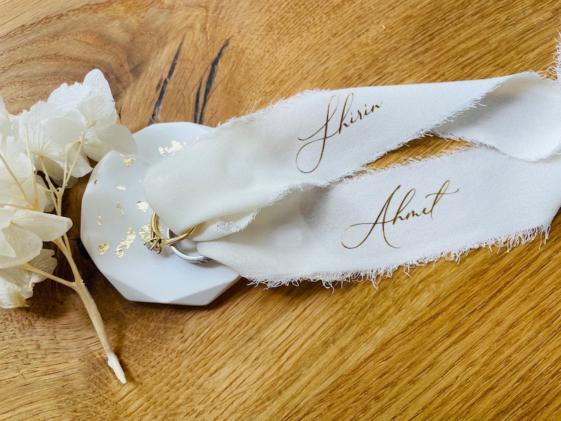 Op de afbeelding: Twee witte stoffen linten met gouden letters, een met de naam "Shirin" en de andere met de naam "Ahmet". De linten liggen op een wit keramiek ringenschotel met gouden vlokken.