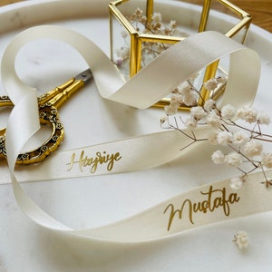 May include: A white satin ribbon with the names "Hayriye" and "Mustafa" written in gold lettering. The ribbon is tied around a small, hexagonal, glass box with a gold frame. The box is filled with dried white flowers.