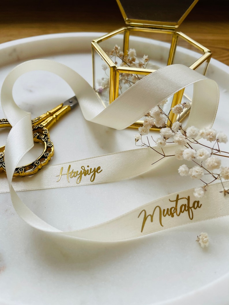 May include: A white satin ribbon with the names "Hayriye" and "Mustafa" written in gold lettering. The ribbon is tied around a small, hexagonal, glass box with a gold frame. The box is filled with dried white flowers.