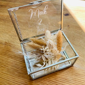 May include: A glass ring box with a silver chain and a lid that says "Maik & Ines". The box is filled with dried flowers.