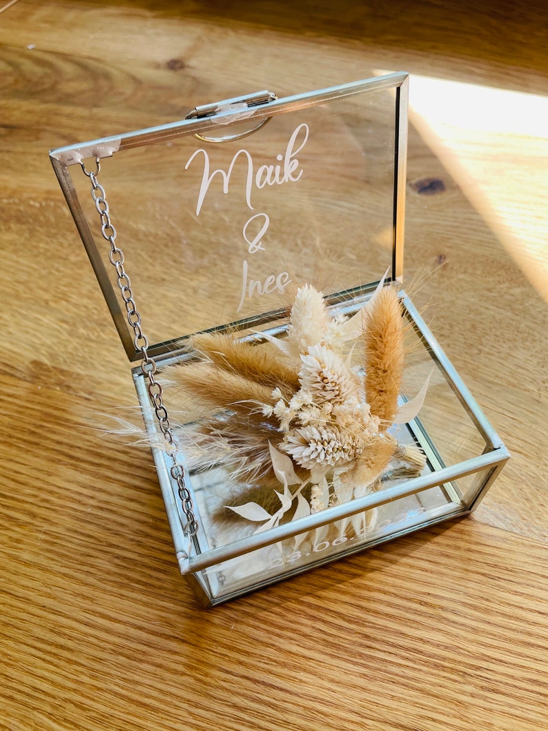 May include: A glass ring box with a silver chain and a lid that says "Maik & Ines". The box is filled with dried flowers.