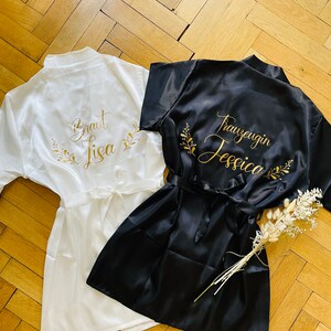 May include: Two satin robes, one white and one black, are displayed on a wooden surface. The white robe has gold text that reads "Braut Lisa" with floral accents. The black robe has gold text that reads "Trauzeugin Jessica" with floral accents.