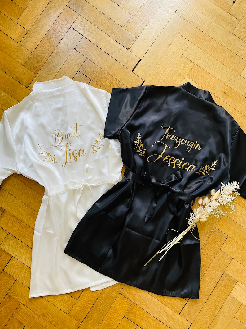 May include: Two satin robes, one white and one black, are displayed on a wooden surface. The white robe has gold text that reads "Braut Lisa" with floral accents. The black robe has gold text that reads "Trauzeugin Jessica" with floral accents.