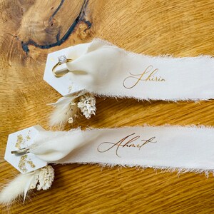 Op de afbeelding: Twee witte zeshoekige plaatskaarten met gouden glitteraccenten en gedroogde bloemen. De bovenste kaart heeft een gouden ringhouder met een diamanten ring. De onderste kaart heeft een gouden clip. Elke kaart heeft een wit lint met gouden kalligrafie. Het bovenste lint zegt "Shirin" en het onderste lint zegt "Ahmet".