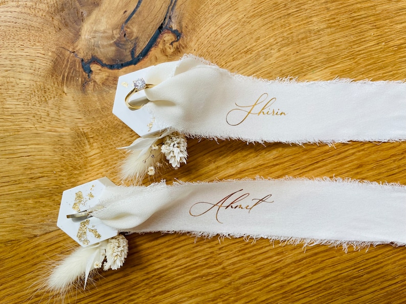 Op de afbeelding: Twee witte zeshoekige plaatskaarten met gouden glitteraccenten en gedroogde bloemen. De bovenste kaart heeft een gouden ringhouder met een diamanten ring. De onderste kaart heeft een gouden clip. Elke kaart heeft een wit lint met gouden kalligrafie. Het bovenste lint zegt "Shirin" en het onderste lint zegt "Ahmet".