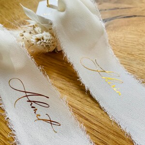 Op de afbeelding: Twee witte satijnen linten met gouden letters. De linten zeggen "Janet" en "Olivia". Op de achtergrond is een ringdoos met een diamanten ring te zien.