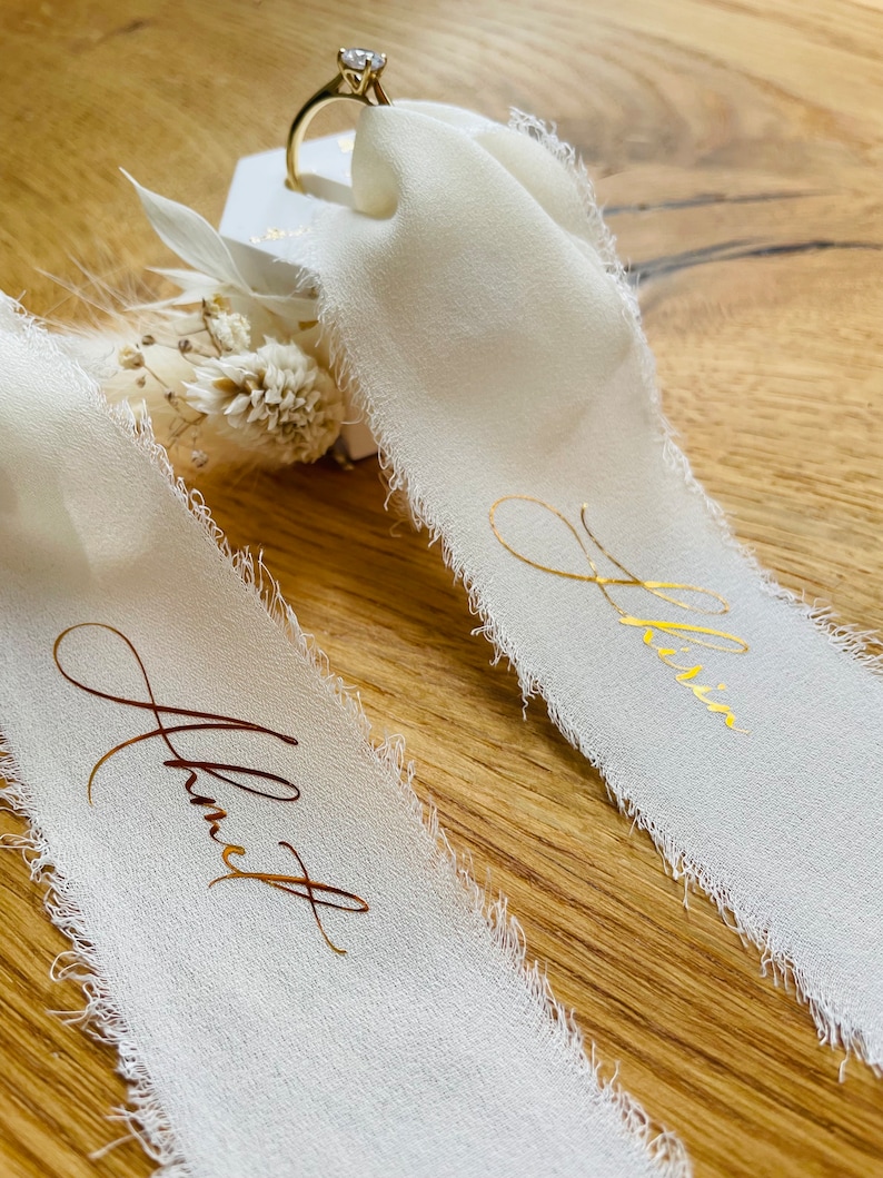 Op de afbeelding: Twee witte satijnen linten met gouden letters. De linten zeggen "Janet" en "Olivia". Op de achtergrond is een ringdoos met een diamanten ring te zien.