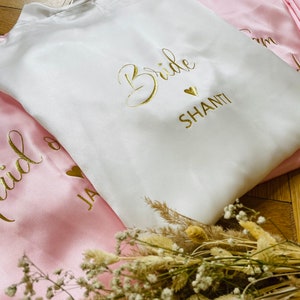 May include: White satin robe with gold text that reads "Bride" and "Shanti". A pink satin robe is visible with gold text. The robes are folded and arranged with dried flowers. Ideal for bridal parties.