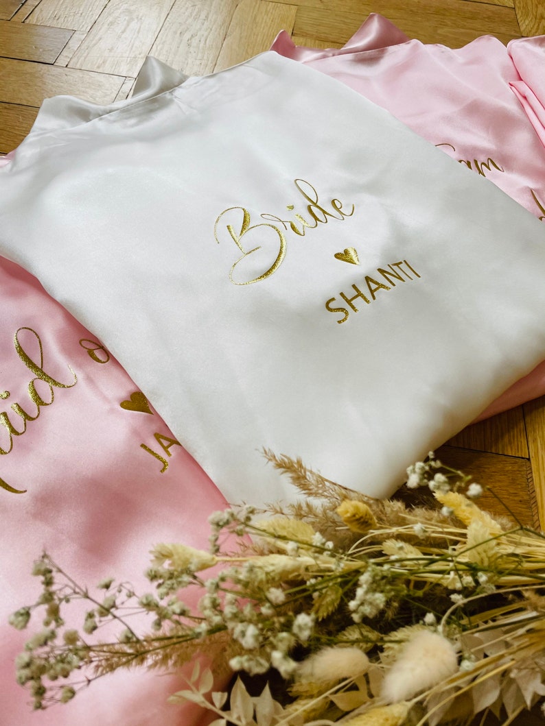 May include: White satin robe with gold text that reads "Bride" and "Shanti". A pink satin robe is visible with gold text. The robes are folded and arranged with dried flowers. Ideal for bridal parties.