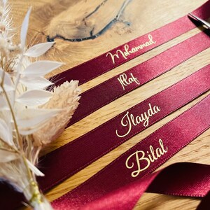 May include: Four burgundy satin ribbons with gold lettering. The names "Mohammad", "Ilaf", "Ilayda", and "Bilal" are written on the ribbons.