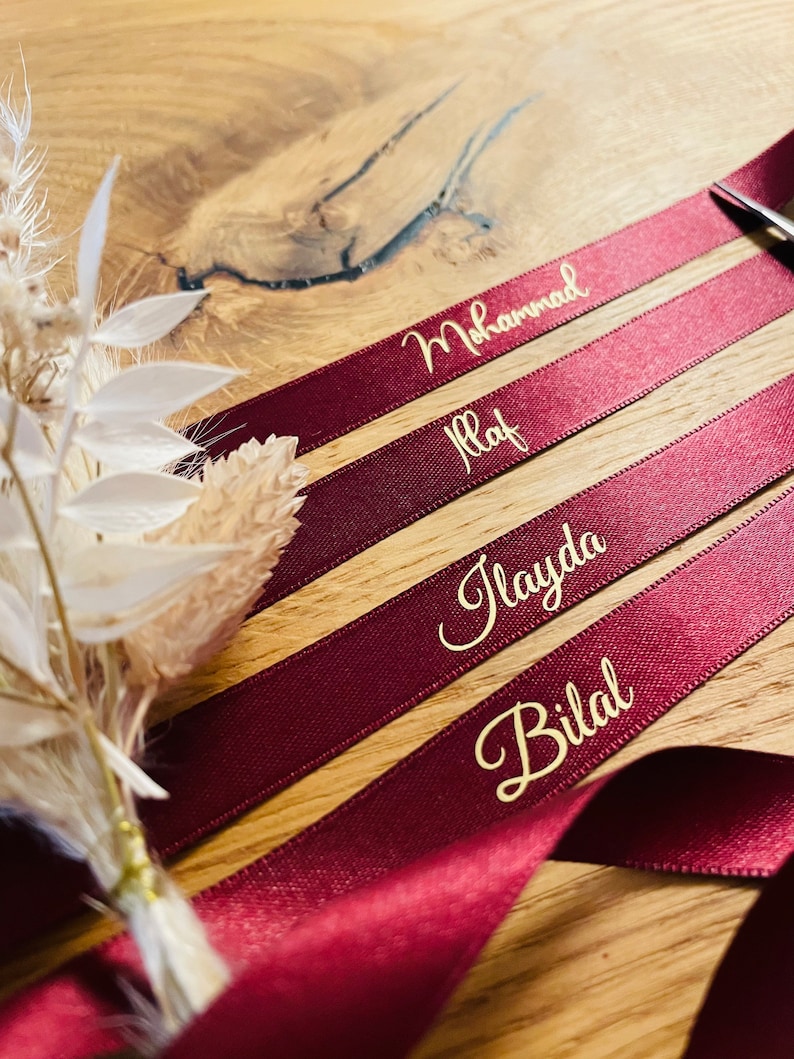 May include: Four burgundy satin ribbons with gold lettering. The names "Mohammad", "Ilaf", "Ilayda", and "Bilal" are written on the ribbons.