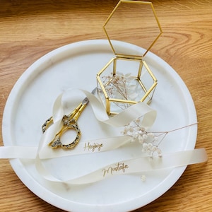 May include: A white marble tray with a gold hexagonal glass box, a white satin ribbon with the names "Haziye" and "Mustafa" written in gold, and a pair of gold scissors.