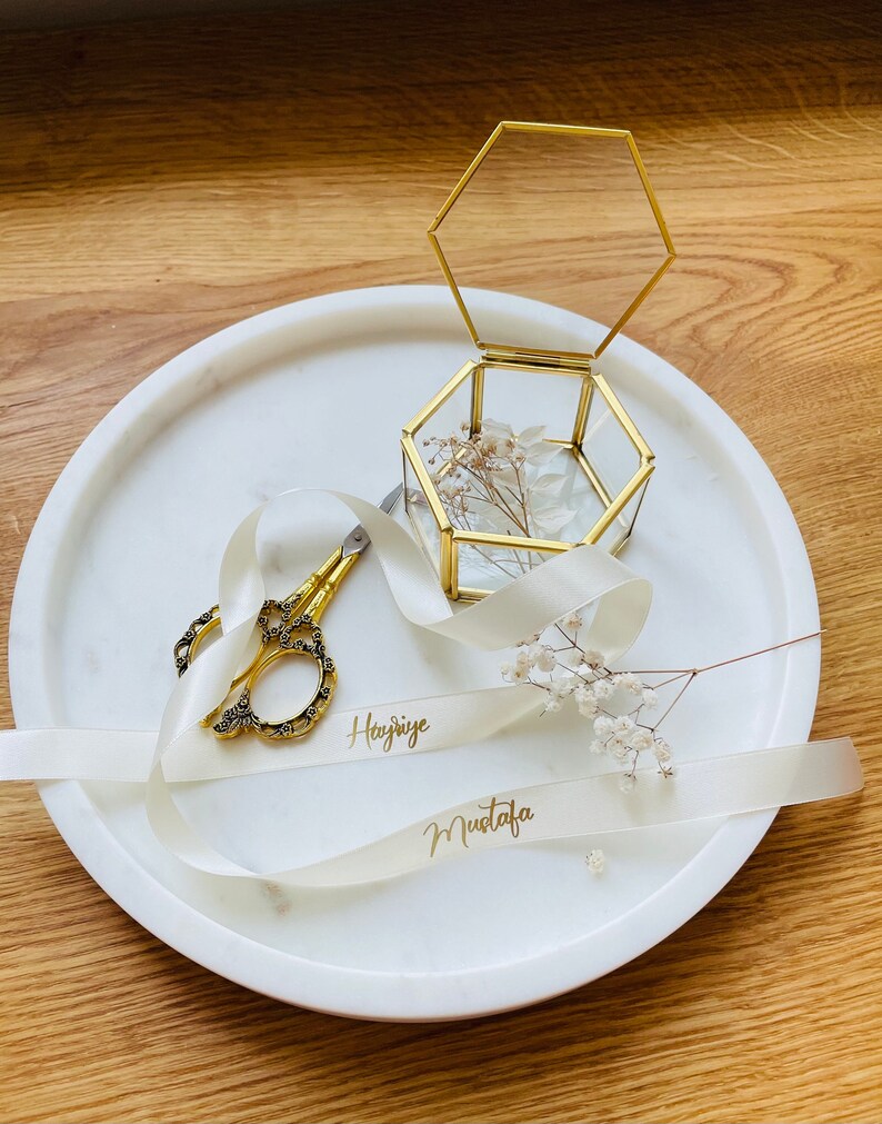 May include: A white marble tray with a gold hexagonal glass box, a white satin ribbon with the names "Haziye" and "Mustafa" written in gold, and a pair of gold scissors.