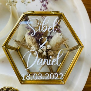 May include: A hexagonal glass ring box with a gold metal frame. The box contains dried flowers and a white inscription that reads "Sibel & Daniel 18.03.2022".