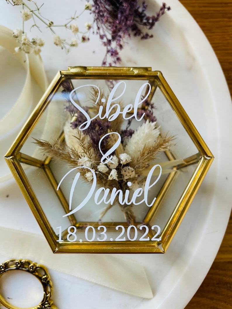 May include: A hexagonal glass ring box with a gold metal frame. The box contains dried flowers and a white inscription that reads "Sibel & Daniel 18.03.2022".