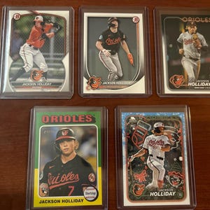 May include: Five baseball trading cards featuring Jackson Holliday of the Baltimore Orioles. The cards are in protective cases. The cards have the Orioles logo and colors, with action shots of the player.