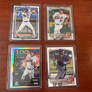 May include: Four baseball trading cards featuring New York Mets players. Each card is encased in a protective sleeve. The cards display images of players in action, with the player's name and team logo visible. The cards are on a wooden surface.