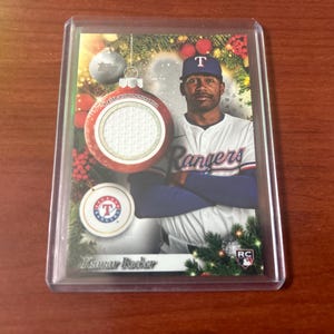 May include: A baseball trading card featuring Kumar Rocker of the Texas Rangers. The card is encased in a protective sleeve and has a festive holiday design with a jersey patch and team logo. The player is wearing a Rangers uniform.