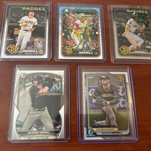 May include: Five baseball trading cards featuring a baseball player in a San Diego Padres uniform. The cards are all in plastic sleeves and have a gold and black design. The player's name is Jackson Merrill.
