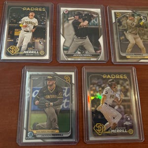 May include: Five baseball trading cards featuring San Diego Padres players, including Jackson Merrill. The cards are in protective sleeves, showcasing action shots and team logos. The cards have the word "PADRES" on them.