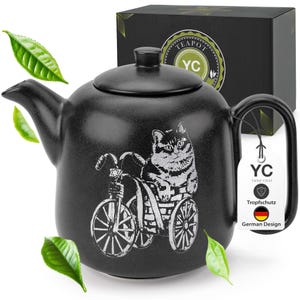 Könnte beinhalten: Schwarze Keramik-Teekanne mit einem Katzen-Tretroller-Design. Die Teekanne hat einen Deckel und einen Griff. Das Design ist in Weiß gehalten. Die Teekanne wird von einer schwarzen Box mit Logo und einem Anhänger mit dem Text "YC Yang Chai" begleitet.