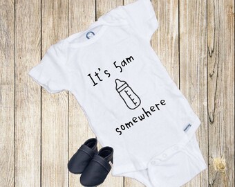 Op de afbeelding: Witte baby romper met zwarte tekst die "Het is 5 uur 's ochtends ergens" luidt en een tekening van een babyfles. Twee zwarte babyschoentjes bevinden zich in de linkerbenedenhoek van de afbeelding.