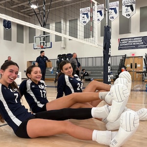 Könnte beinhalten: Drei junge Frauen in marineblauen Volleyball-Trikots mit weißen Akzenten sitzen auf einem Holzboden. Sie lächeln und tragen weiße Knieschoner. Volleyballbälle liegen in der Nähe. Ein Volleyballnetz und Banner sind im Hintergrund.