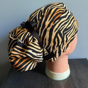 May include: A scrub cap featuring a tiger stripe pattern in shades of orange, black, and cream. The cap has a gathered back section designed to accommodate hair, secured with a black ribbon tie that forms a small bow at the nape of the neck.