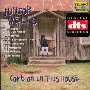 Puede incluir: Portada del álbum "Come On In This House" de Junior Wells. La imagen muestra a un hombre sentado en un porche, con el título del álbum y el nombre del artista en letras grandes. La portada también enumera a los guitarristas invitados y presenta un logotipo "Digital DTS Surround".