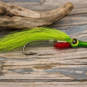 Puede incluir: Un señuelo de pesca con una larga cola de plumas de color chartreuse brillante, un anzuelo de metal plateado y un cuerpo cilíndrico verde. El señuelo incluye un ojo con borde dorado y pupila negra, además de fibras rojas en el anzuelo.