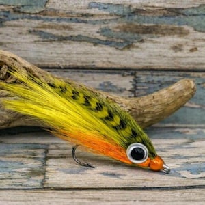 Puede incluir: Un señuelo de pesca con plumas de color amarillo brillante y naranja con patrones de chevrón negros en la parte superior. El señuelo tiene un ojo plateado y negro prominente en el lateral, una cabeza naranja y un anzuelo de metal curvado.