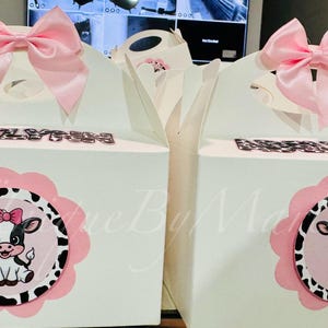 May include: Two white gift boxes with pink bows and cow-themed decorations. Each box features a pink and black cow illustration and the name "ABIGAIL" in silver letters. The boxes are designed for a party or celebration.