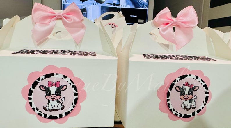 May include: Two white gift boxes with pink bows and cow-themed decorations. Each box features a pink and black cow illustration and the name "ABIGAIL" in silver letters. The boxes are designed for a party or celebration.