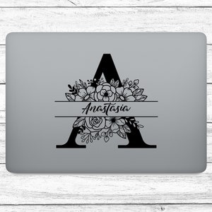 Könnte beinhalten: Ein schwarzes florales Monogramm mit dem Buchstaben "A" und dem Namen "Anastasia" in Schreibschrift. Der Buchstabe ist mit floralen und Blattmotiven verziert. Das Design befindet sich auf einem grauen Laptop.