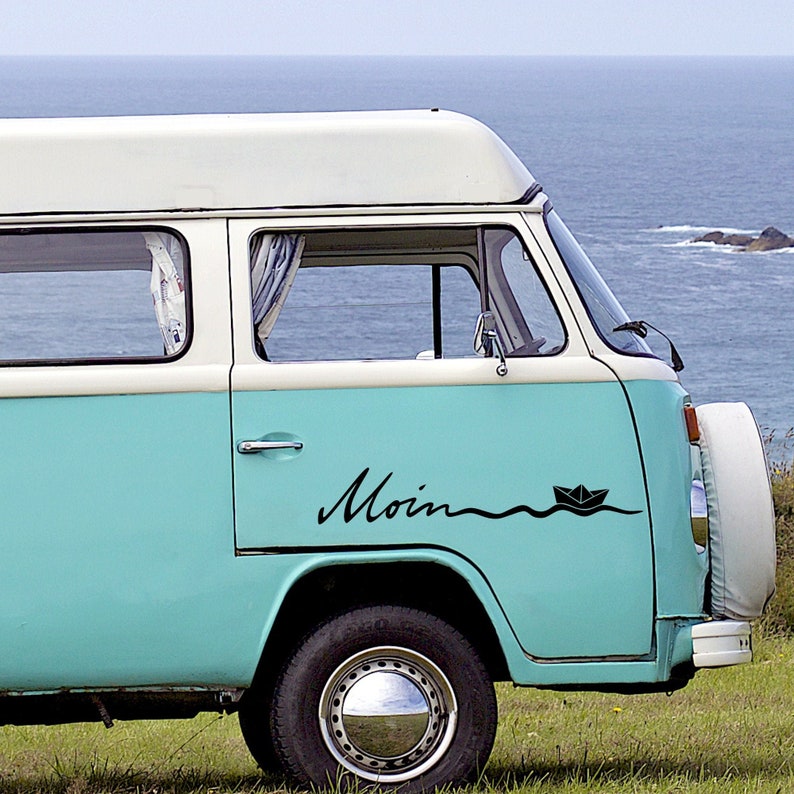 May include: A vintage teal and white camper van with the word "Moin" in black script and a paper boat graphic. The van is parked on a grassy hill with the ocean in the background.